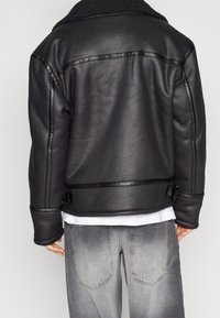 Sort læderjakke med en krave af lammeskind, der har tekstureret materiale, spænde-detaljer og kontrasterende sort syning langs sømmene.