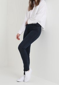 Persona con jeans ajustados oscuros, camisa blanca de botones oversize y zapatillas blancas, de pie contra un fondo claro y sencillo.