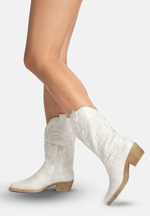 Ben iført hvide cowboystøvler af læder til midt på læggen med broderede blomsterstrukturer og træhæle, den ene støvle løftet på tåen.