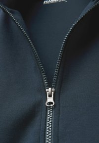 Chaqueta de cremallera azul marino que presenta una cremallera de metal plateada con una lengüeta texturizada, tela suave y una vista en primer plano del área de la cremallera.