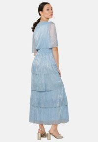 Jasnoniebieska sukienka warstwowa z przezroczystymi krótkimi rękawami, z połyskującą strukturą i dopasowaną talią. Założona z srebrnymi sandałami na cienkich obcasach.