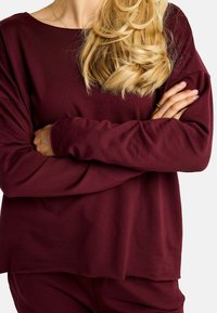 Bordo majica z dolgimi rokavi, širokim izrezom in sproščenim krojem. Mehka tkanina z gladko teksturo, s rahlo spuščenimi rameni.