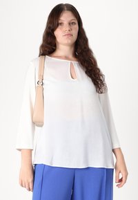 Witte, lichte top met een ronde hals en een keyhole-detail; gecombineerd met blauwe wijde broek en een beige schoudertas.