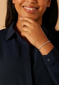 Zilveren armband met kleine heldere steentjes, gedragen op een donkerblauw shirt met lange mouwen. Gladde textuur, eenvoudig ontwerp, close-up van hand en gezicht.