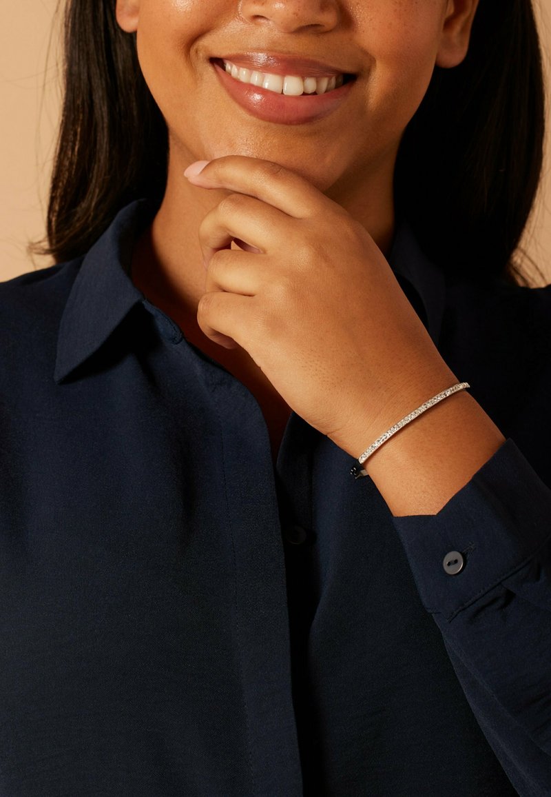 Zilveren armband met kleine heldere steentjes, gedragen op een donkerblauw shirt met lange mouwen. Gladde textuur, eenvoudig ontwerp, close-up van hand en gezicht.