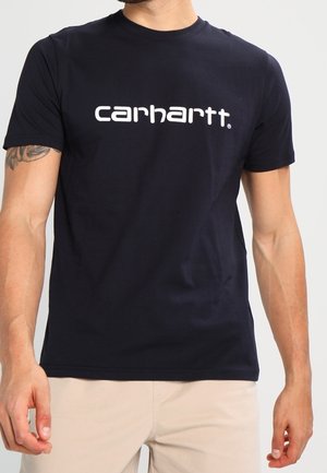 Man draagt een zwart Carhartt T-shirt en beige broek, met een tatoeage zichtbaar op zijn linker bovenarm.
