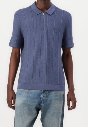 Mann trägt ein kurzärmeliges blaues Strick-Poloshirt mit Reißverschluss vorne und hellblaue Jeans auf weißem Hintergrund.