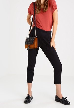 Femme portant un haut couleur rouille, un pantalon noir court, des mocassins noirs et un sac à main marron avec un rabat à motif noir et une bandoulière.