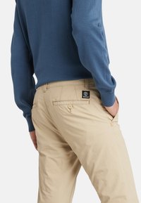Chinos beige con una texture liscia, tasche posteriori con bottoni e un piccolo etichetta del logo Timberland sul lato destro. Maglione blu visibile.