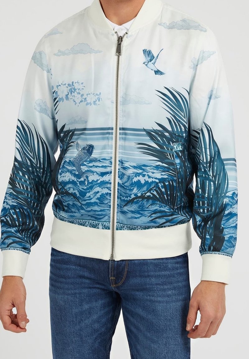 Homme portant une veste blanche zippée ornée de vagues bleues, de poissons, de feuilles de palmier et de motifs d'oiseaux, associée à un jean bleu.