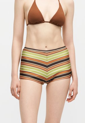 Femeie care poartă un costum de baie maro și pantaloni scurți cu talie înaltă, drepți cu model chevron verde, portocaliu, negru și alb, stând pe un fundal simplu.