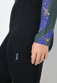 Czarne dopasowane legginsy o gładkiej fakturze i kontrastowym logo. Góra ma kolorowy abstrakcyjny wzór w odcieniach zieleni, fioletu i złota.