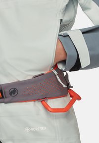 Person iført en lysegrå Gore-Tex-jakke og en grå klatresele med orange detaljer samt en fastgjort orange karabiner.