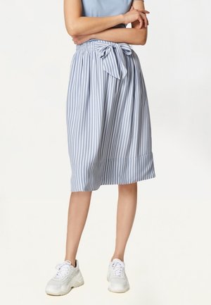Vrouw die een midi-rok draagt met blauw-witte verticale strepen en een strik in de taille, witte sneakers en een lichtblauw topje, armen over elkaar.