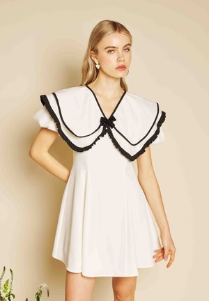 Vrouw in een witte korte jurk met grote kraag, zwarte bies en strik, staand tegen een beige achtergrond met haar hand op haar heup.