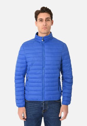 Uomo con giacca trapuntata blu brillante con zip e collo alto e jeans blu, in piedi davanti a uno sfondo bianco semplice.