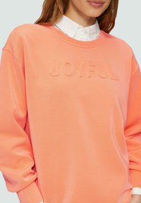 Koraal kleurige sweatshirt met een geperst "JOYFUL" ontwerp, ribbelachtige ronde hals en een relaxte pasvorm. Lange mouwen met gatherings aan de manchetten.
