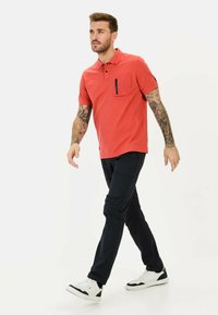 Rode polo met een rits op de borst, gecombineerd met zwarte broeken en witte sneakers. Model heeft tatoeages en een neutrale pose.