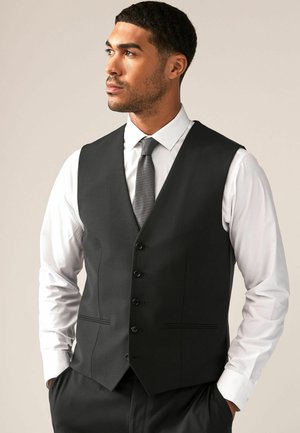 Chaleco negro de tela suave, con escote en V profundo, seis botones en la parte delantera y dos bolsillos delanteros. Usado sobre una camisa blanca.