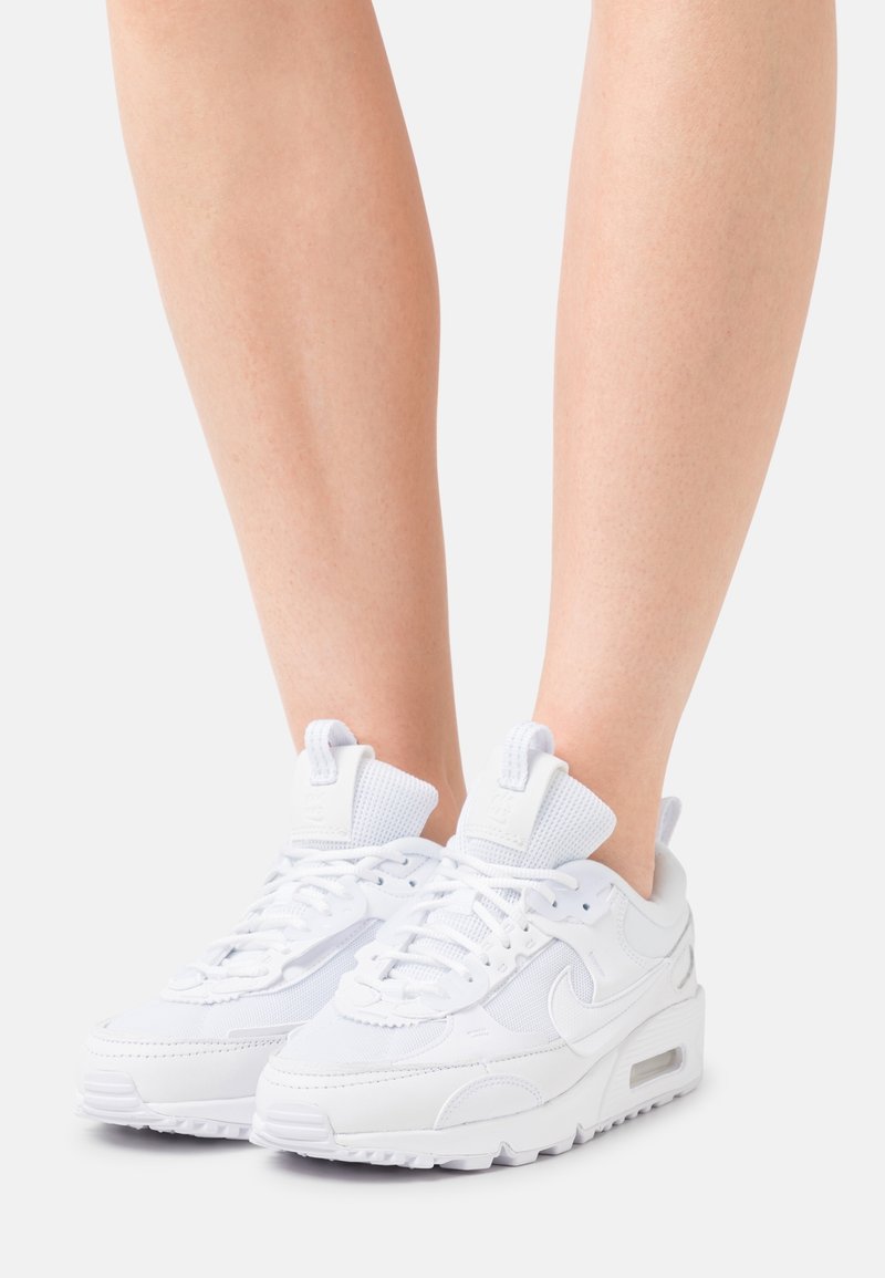 Personne portant des baskets Nike blanches à lacets, debout sur un fond clair et uni ; seules les jambes et les chaussures sont visibles.