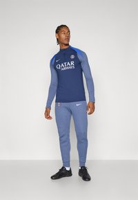 Marineblå langærmet atletisk top med logo og blå skulderaksenter, parret med lyseblå tapered joggerbukser og sorte træningssko.
