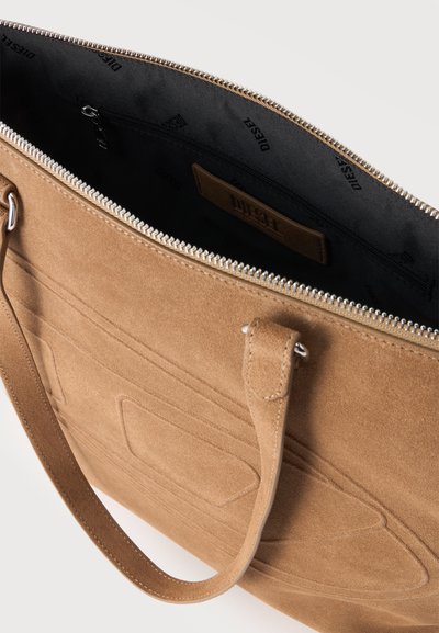 Sac cabas en daim camel avec logo embossé, fermeture éclair argentée et doublure intérieure noire avec poche zippée et étiquette de marque en tissu.
