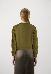 Sudadera corta color verde olivo con mangas raglán y bolsillos laterales, combinada con una falda gris de longitud media con detalle de cremallera.
