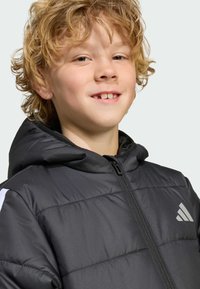 Veste à capuche noire rembourrée avec zip, présentant des accents blancs sur les manches et un logo gris sur le haut à gauche de la poitrine.