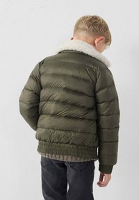 Giacca puffer verde oliva con colletto in shearling bianco, design trapuntato, polsini e orlo elasticizzati, indossata sopra un maglione grigio e jeans neri.
