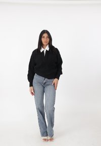 Maglione nero a trecce con scollo a V, abbinato a jeans azzurri a vita alta e tacchi bianchi aperti. Camicia bianca solida con colletto sotto.