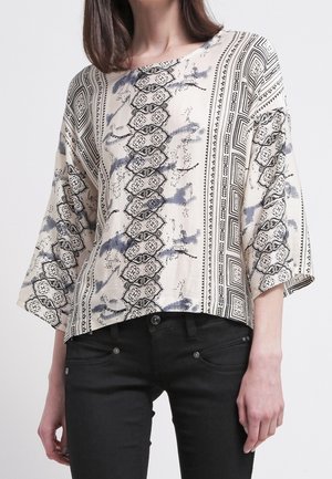 Haut crème à motif floral avec des accents géométriques noirs, coupe loose et manches 3/4. Associé à un pantalon noir, le tissu semble doux et texturé.