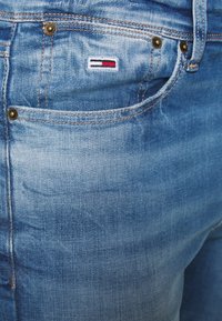 Vaqueros de mezclilla azul claro con un acabado desgastado, que cuentan con dos bolsillos delanteros, un pequeño logo bordado en rojo y blanco, y herrajes de bronce.