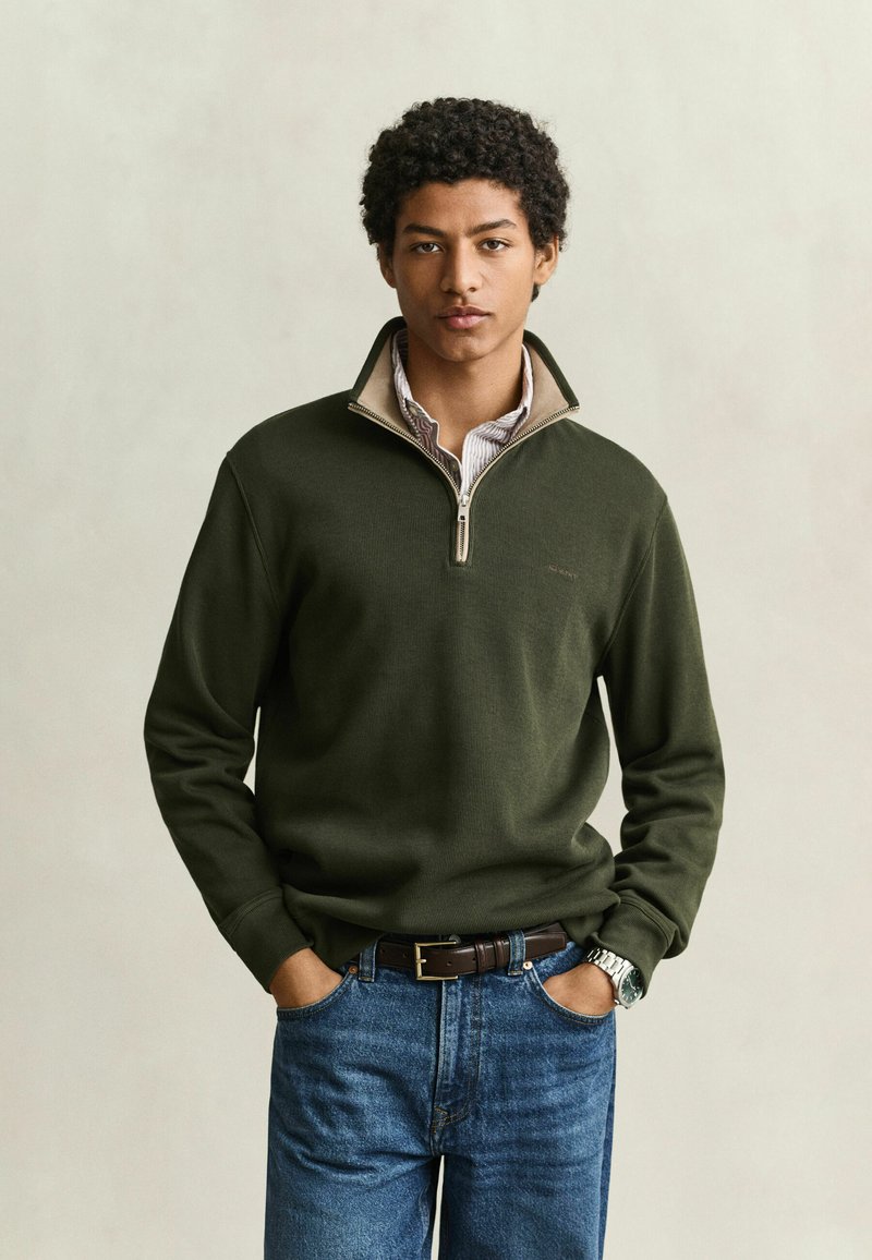 Jeune homme aux cheveux bouclés portant un pull vert à fermeture quart de zip, une chemise rayée en dessous, un jean bleu, une ceinture marron et une montre argentée au poignet, debout avec les mains dans les poches.