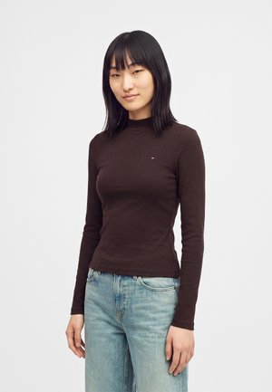 Junge Frau mit glatten schwarzen Haaren, die ein enganliegendes braunes Langarmoberteil und hellblaue Jeans vor einem schlichten Hintergrund trägt.