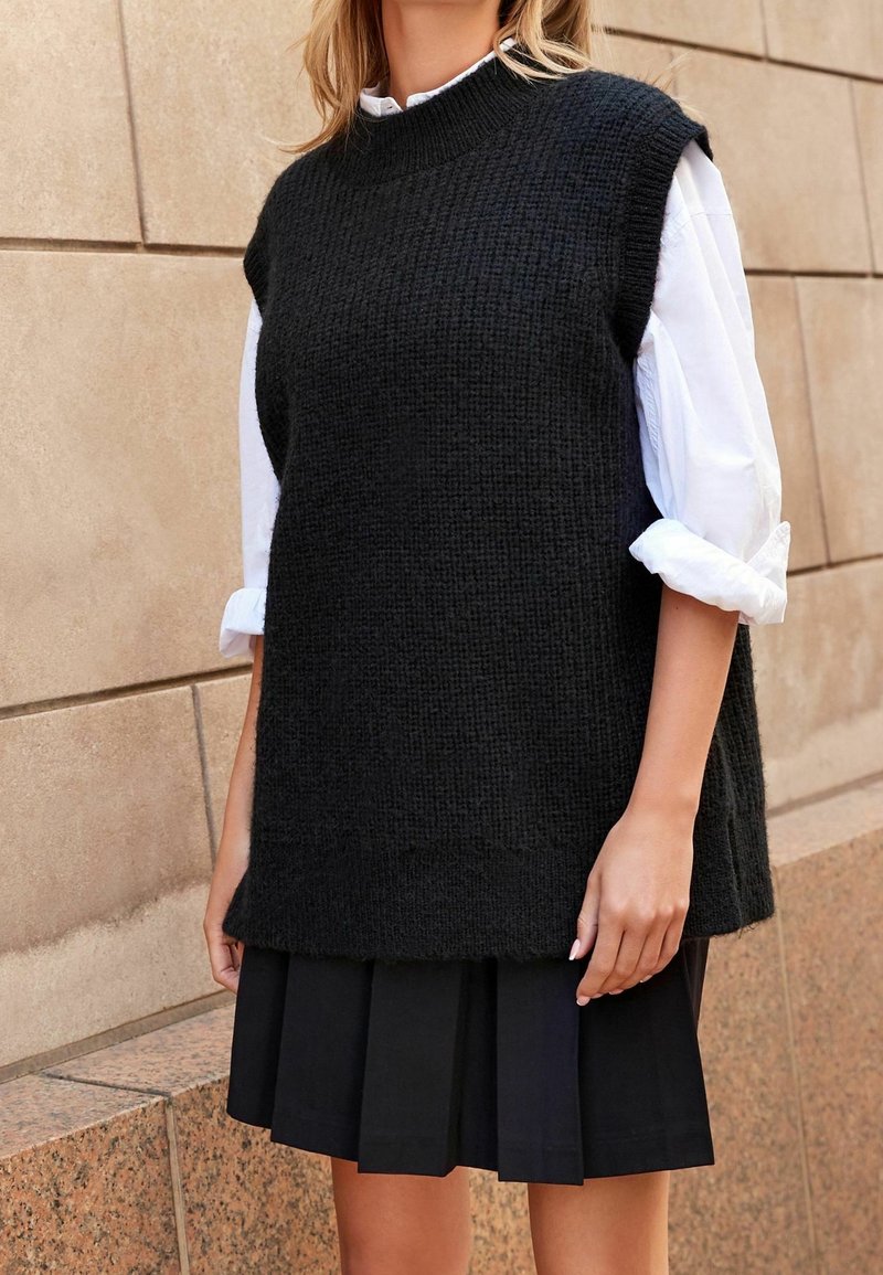 Femme portant un gilet sans manches en tricot noir sur une chemise blanche retroussée et une jupe plissée noire, debout contre un mur beige carrelé.