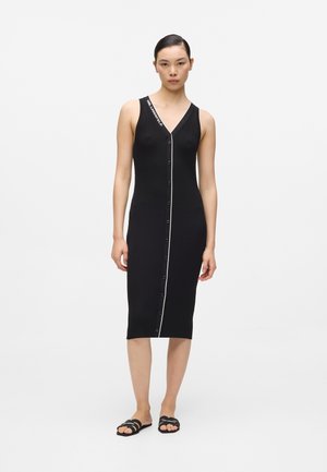 Kobieta ubrana w czarną, bezrękawnikową, prążkowaną sukienkę midi z przodu zapinaną na guziki i z białą lamówką, w połączeniu z czarnymi klapkami.