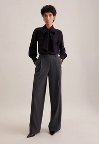 Blusa nera con scollo a fiocco, abbinata a pantaloni grigi a vita alta e gamba larga. Tessuto liscio e vestibilità su misura in ogni dettaglio.