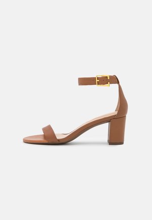 Brązowy sandał na słupku z paskiem wokół kostki i złotą klamrą, odkrytymi palcami i niskim obcasem na białym tle.