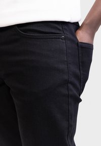 Άτομο που φοράει μαύρο παντελόνι με το δεξί χέρι στην μπροστινή τσέπη, λευκό πουκάμισο ορατό στη μέση, απέναντι σε απλό λευκό φόντο.