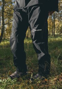 Čierne vonkajšie nohavice s vreckom na zips, s logom na stehne, vyrobené z odolného materiálu, v pozadí s lesným prostredím.