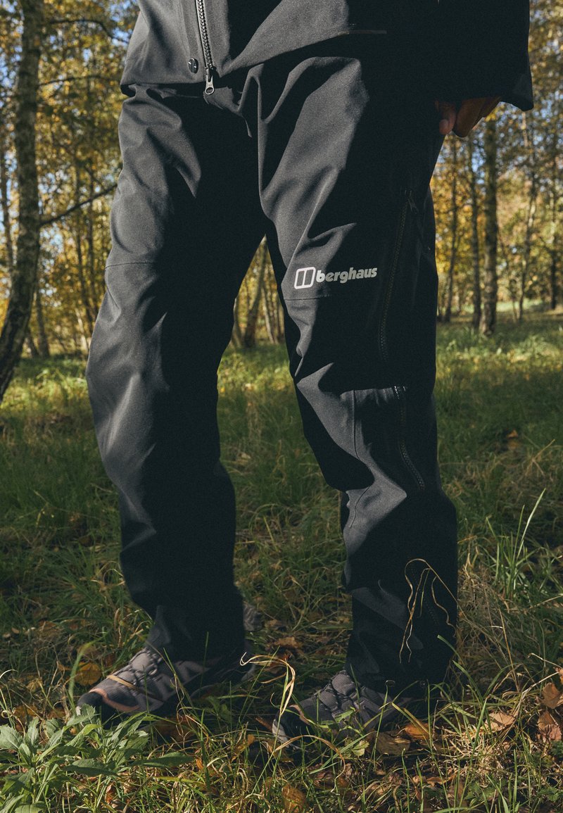 Čierne vonkajšie nohavice s vreckom na zips, s logom na stehne, vyrobené z odolného materiálu, v pozadí s lesným prostredím.
