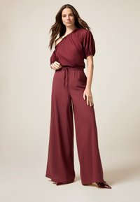 Bordeaux off-shoulder jumpsuit met pofmouwen, geplooide taille en wijd uitlopende broekspijpen. Glad materiaal met een subtiele glans, gecombineerd met puntige schoenen.