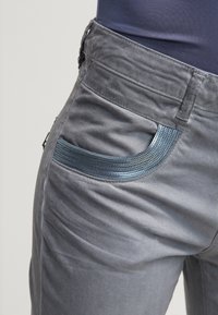 Šedé kalhoty s texturovaným modrým akcentem na kapse. Tkanina vypadá hladce a design zahrnuje detail přední kapsy.