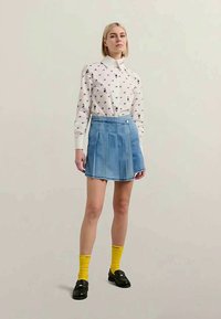 Bela srajca z majhnimi pisanimi vzorci, svetlo modra denimska krila, nogavice do kolen v rumeni barvi in črne čevlje. Preprost, sproščen stil.