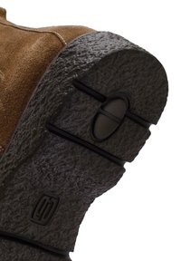 Bota de ante marrón con una suela de goma negra texturizada, que presenta surcos distintivos y un logo circular en el talón para proporcionar tracción y soporte.