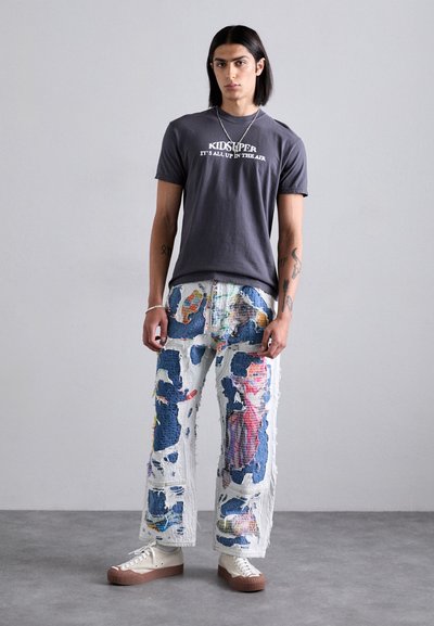 Gray graphic t-shirt with "KID SUPER" text, paired with loose, distressed denim pants featuring a colorful abstract pattern. White sneakers with brown soles.