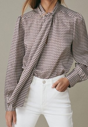 Femme portant un chemisier à manches longues à motifs avec un nœud à l'avant et un pantalon blanc taille haute, une main dans la poche.