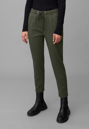 Personne portant un pantalon à cordon de serrage vert olive et des bottes noires à tige haute, les mains relâchées sur les côtés, sur un fond gris uni.