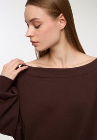 Felpa oversize marrone scuro con scollatura asymmetrica, polsini a costine e tessuto morbido e testurizzato. Delicati dettagli delle cuciture sulla scollatura.
