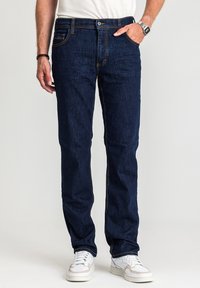 Ciemnoniebieskie jeansy z denimu o prostym kroju nogawek, z �żółtym przeszyciem i przednimi kieszeniami. Noszone z białymi tenisówkami i prostą białą koszulą.